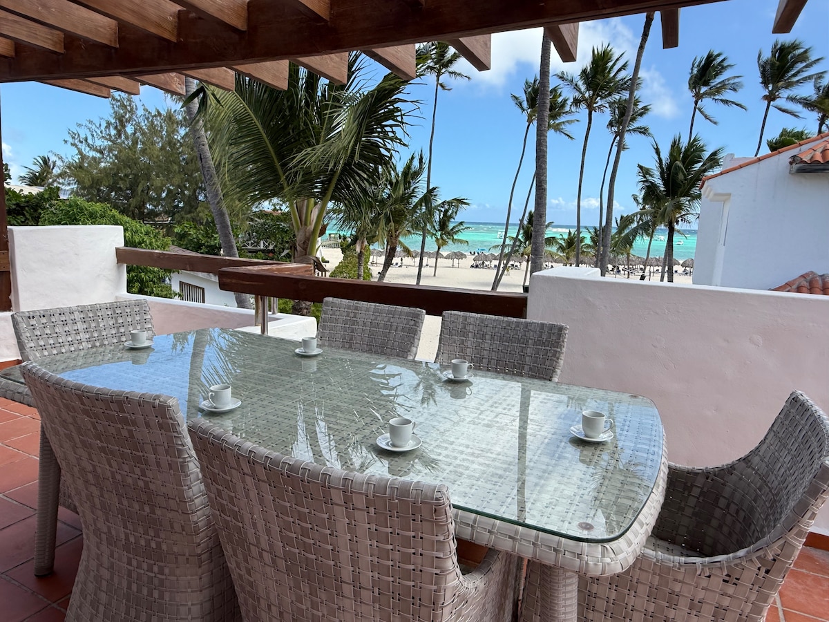 A dining area features a glass-top table surrounded by six wicker chairs, with coffee cups placed neatly on the table. A view of the beach and palm trees is visible in the background, providing a bright and open atmosphere.