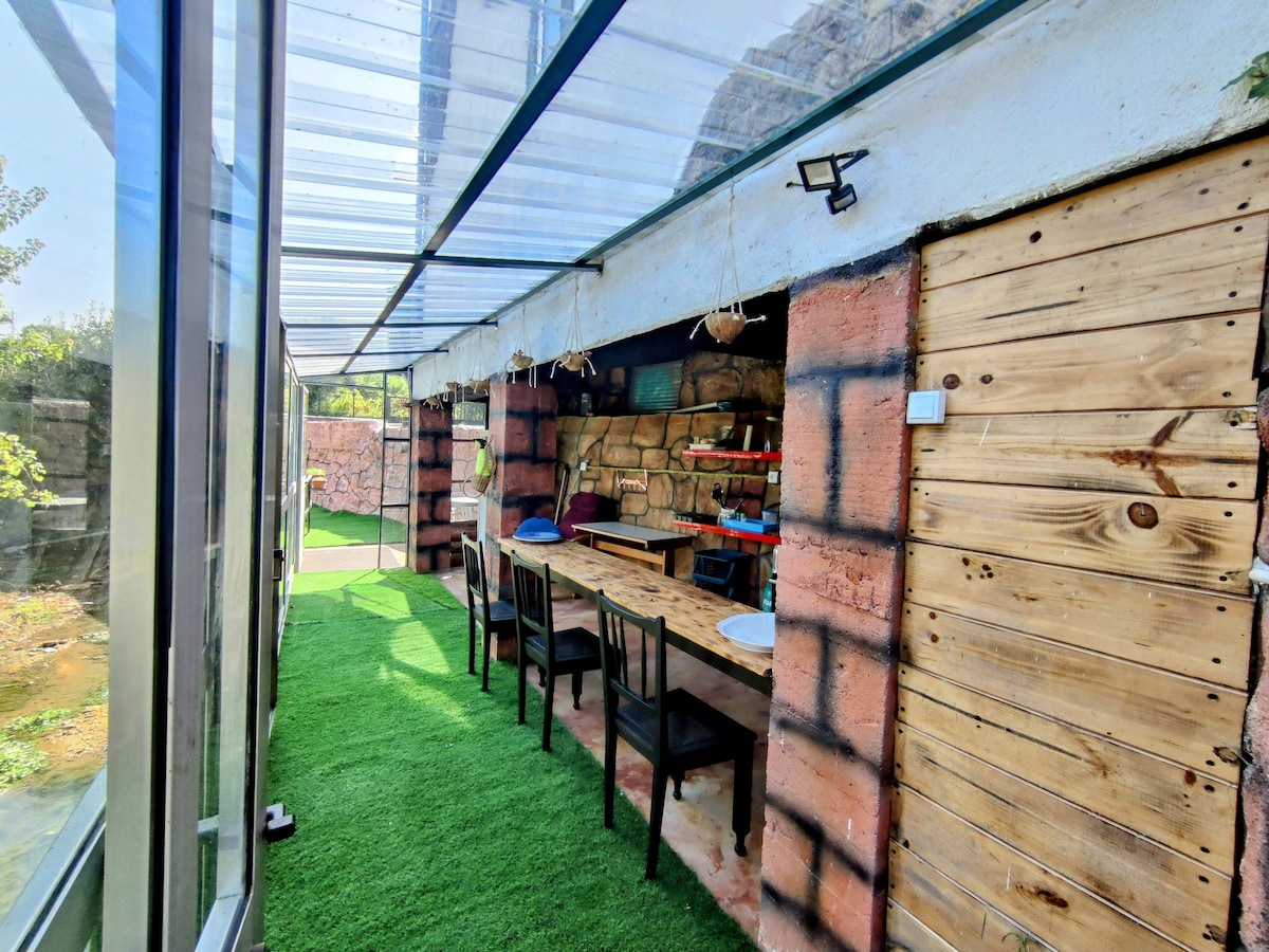 A sheltered outdoor area is shown, featuring synthetic grass flooring and natural stone walls. Wooden tables are arranged alongside black chairs, creating a casual dining space. Sunlight filters through the glass roof, illuminating the inviting setting, with greenery visible outside.