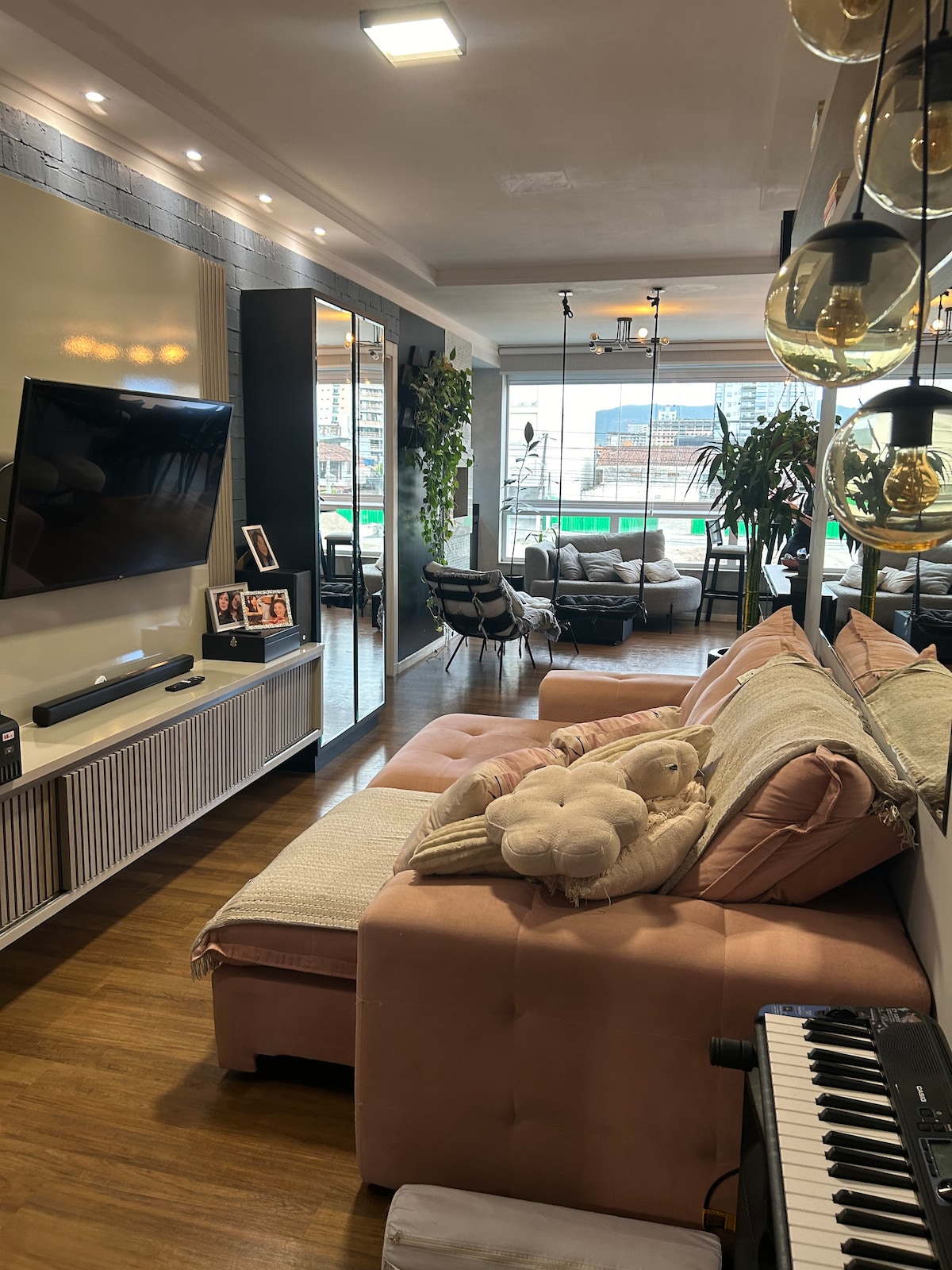A spacious living area features a large pink sectional sofa adorned with plush pillows and a stuffed toy. A flat-screen television is mounted on the wall, and large windows allow natural light to fill the room, while greenery adds a fresh touch.