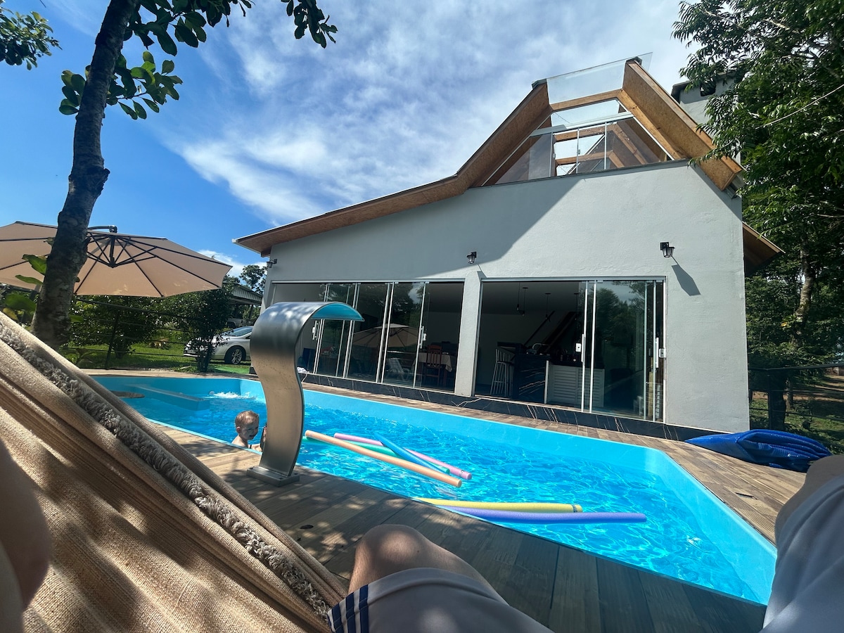 A spacious outdoor area is highlighted by a private pool featuring a gentle fountain and surrounded by wooden decking. Large glass doors provide a view of the interior living space, while sun loungers and a shaded umbrella are positioned nearby, creating a welcoming space for relaxation.