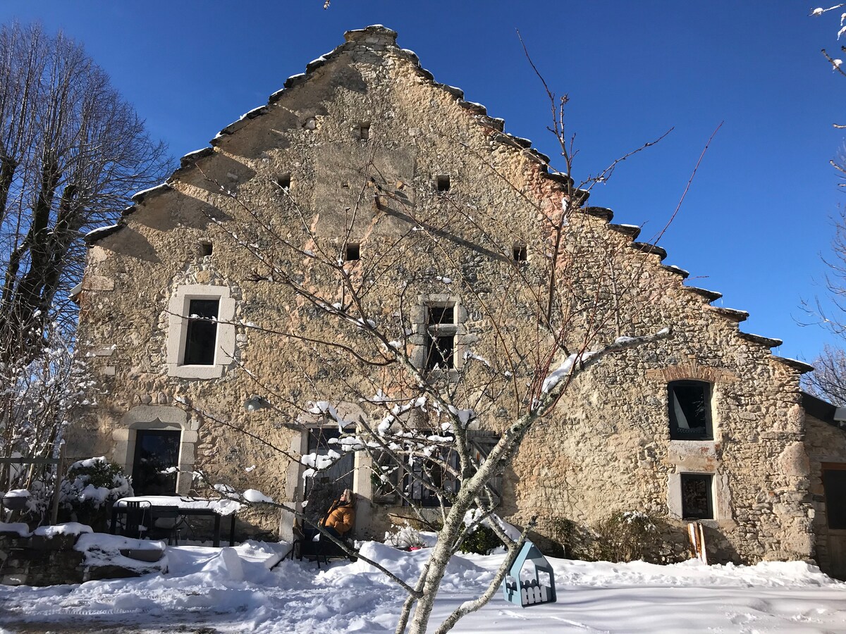 The exterior of a renovated stone house is displayed, framed by snow-covered ground. The structure features a sloped roof with traditional tiles and several windows, allowing natural light. A small garden area is visible in the foreground, with bare trees adding to the winter scenery.