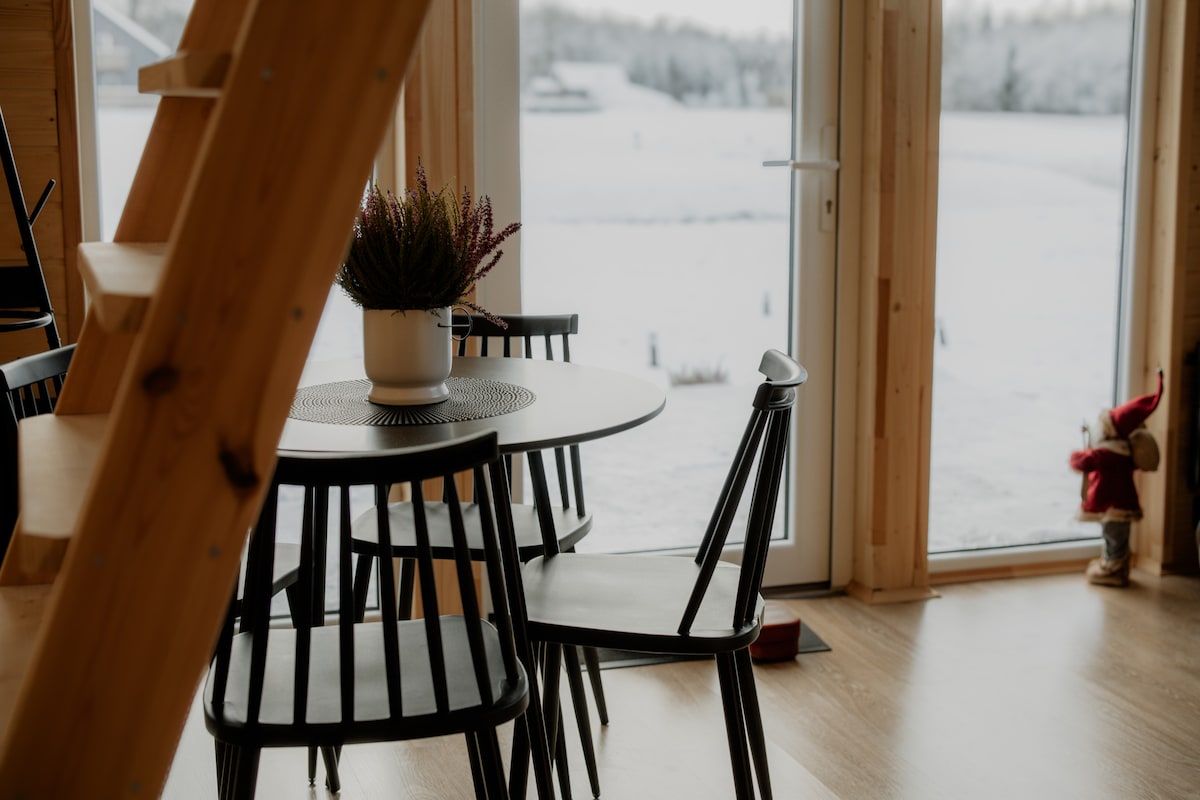 A dining area is presented with a round table surrounded by six black chairs. A potted plant sits at the center of the table. Large glass doors allow views of a snowy landscape, enhancing the connection between indoor and outdoor spaces.