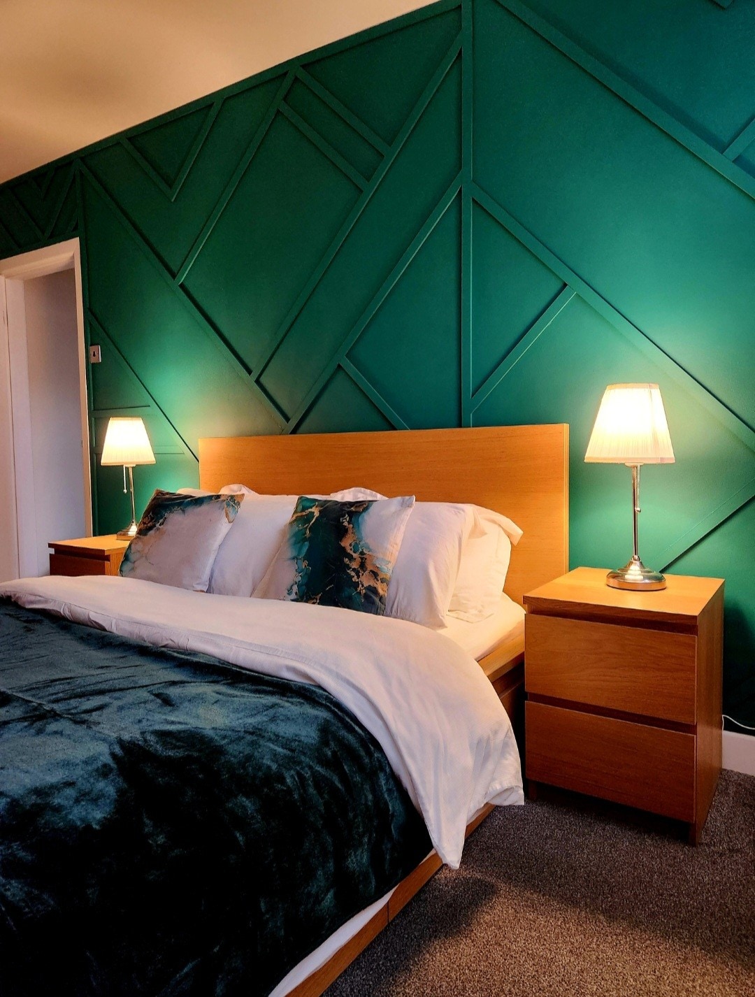 A spacious bedroom features a wooden headboard and nightstands, accented by teal green geometric wall panels. Soft pillows and a cozy spread adorn the bed. Two bedside lamps provide gentle illumination, enhancing the inviting atmosphere.