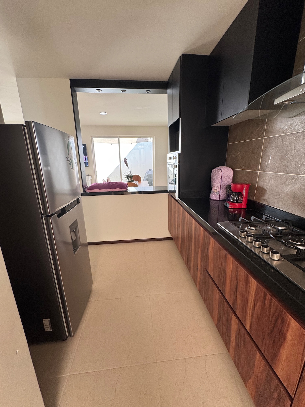 The kitchen features sleek dark cabinetry and a contrasting light countertop. A stainless steel refrigerator and modern gas stove are visible, while a window offers a view into the adjoining space. Natural light enhances the clean and organized environment.