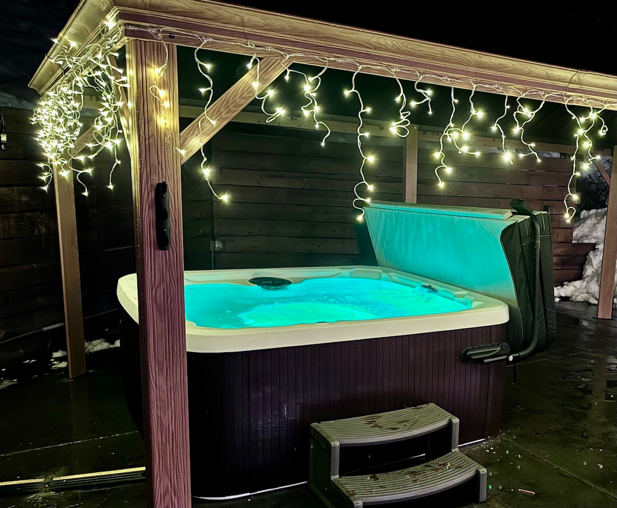 A hot tub sits beneath a wooden gazebo adorned with soft fairy lights, creating a serene ambiance. The tub is filled with inviting water, and a set of steps leads up to it. Snow can be seen on the surrounding ground, indicating a cozy retreat.