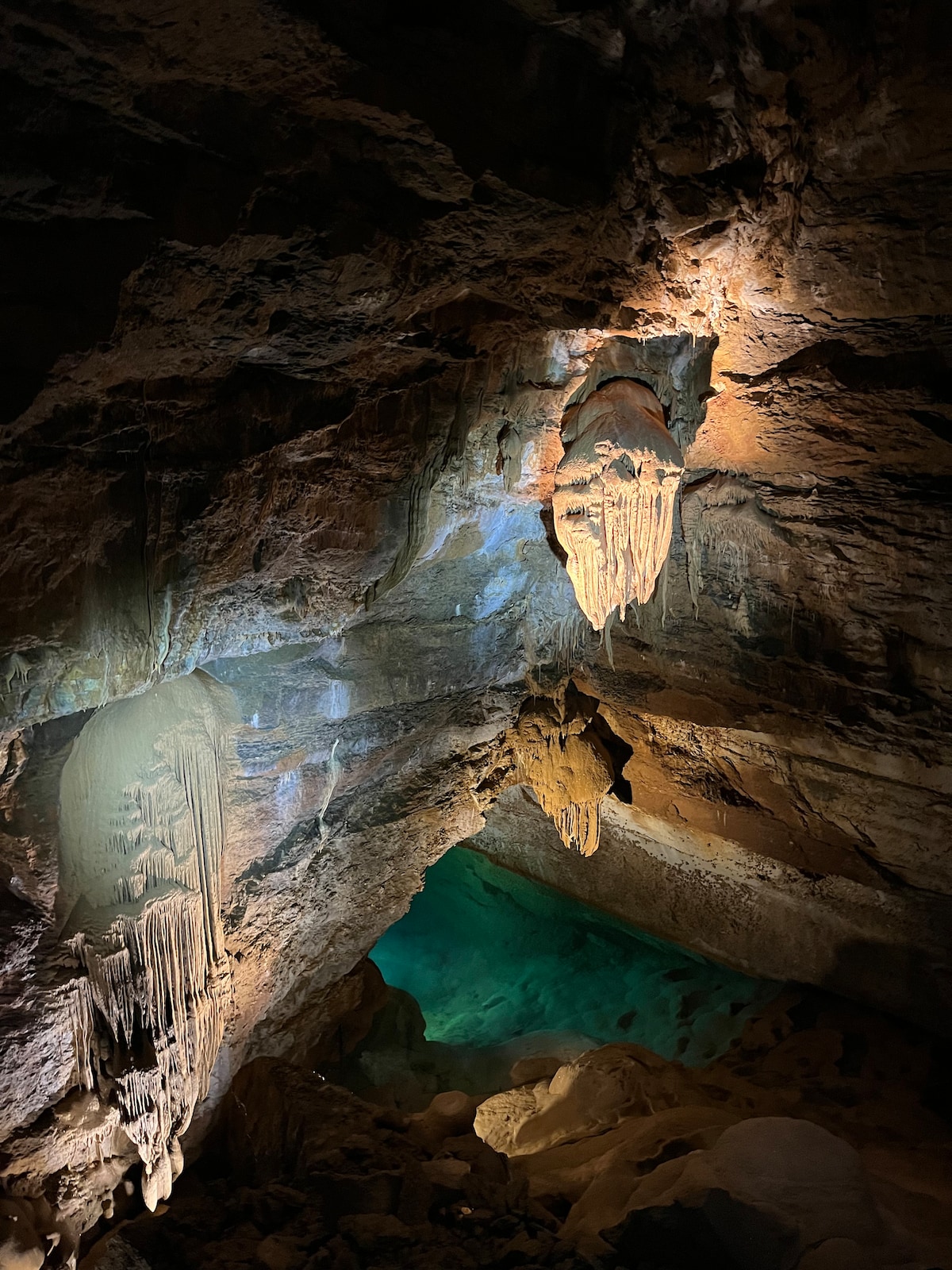 An underground cave is illuminated by subtle lighting, revealing intricate rock formations and mineral deposits. Stalactites cascade from the ceiling, while a pool of deep blue water can be seen at the cave's base, enhancing the natural beauty of the rugged surroundings.