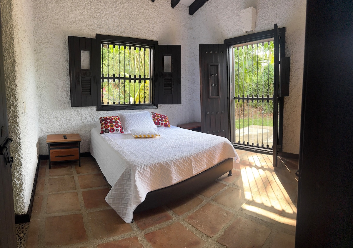 A spacious bedroom is presented with a large bed featuring a white quilt and colorful accent pillows. Natural light filters through the open windows, illuminating the terracotta tile floor. A wooden nightstand is positioned beside the bed, enhancing the room's functional layout.