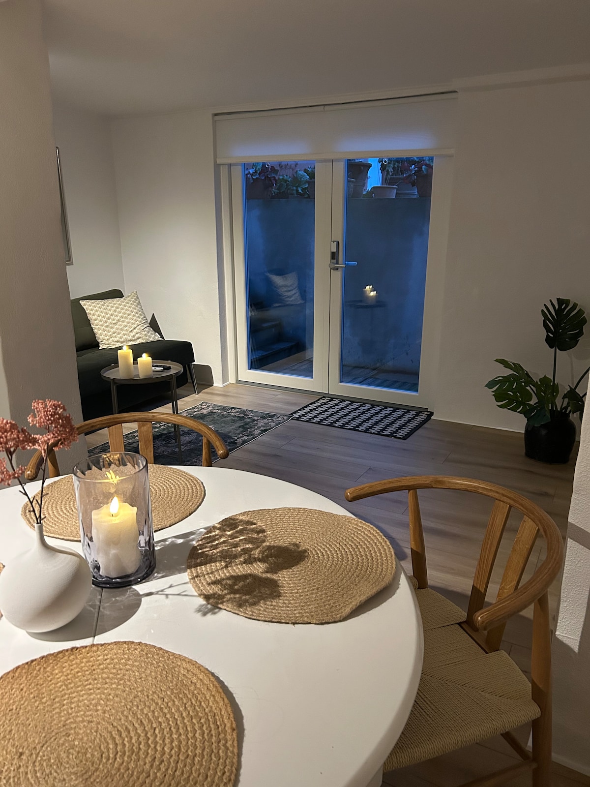 A dining area features a round table set with woven placemats and a decorative vase. A cozy seating area with a dark couch provides a relaxed atmosphere. Light from candles illuminates the space, while glass doors open to a small outdoor area with greenery.