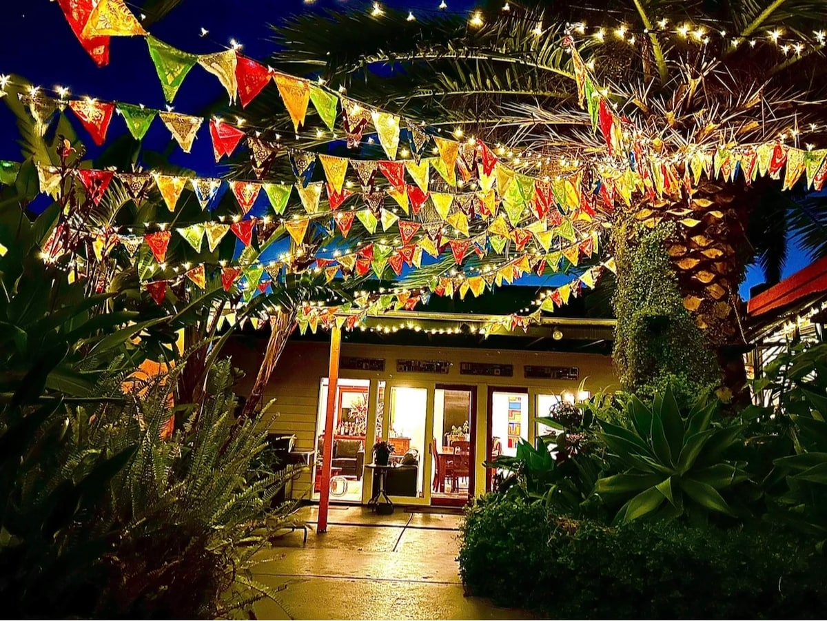 Colorful bunting and warm string lights hang from trees, creating a festive atmosphere in the garden at night. The soft glow illuminates the pathway leading to large glass doors, providing a view into the inviting living space of the bungalow.