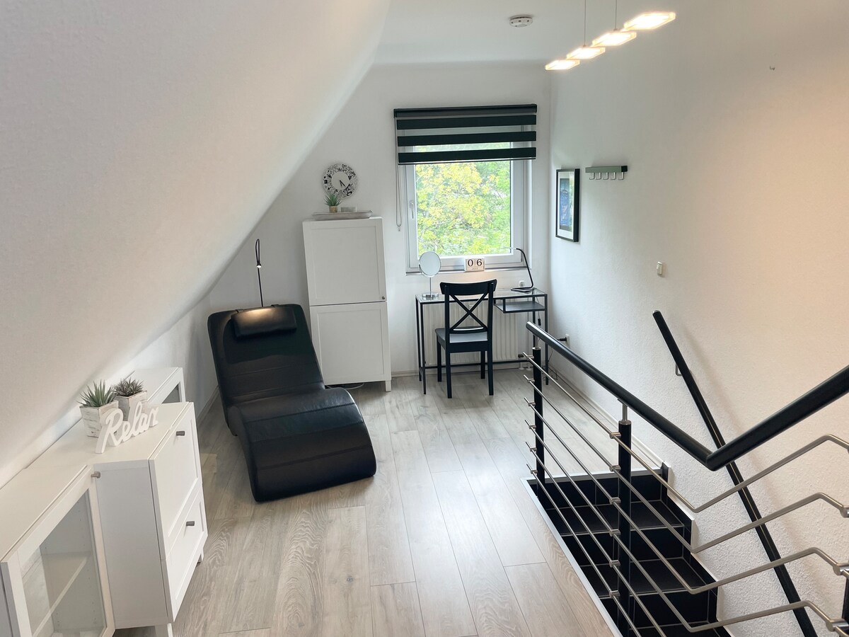 A bright and inviting corner of the upper level features a relaxing chaise lounge in black, positioned beside a window with a view of greenery. A simple desk and chair offer a workspace, while a small white cabinet adds storage. Soft lighting illuminates the space.