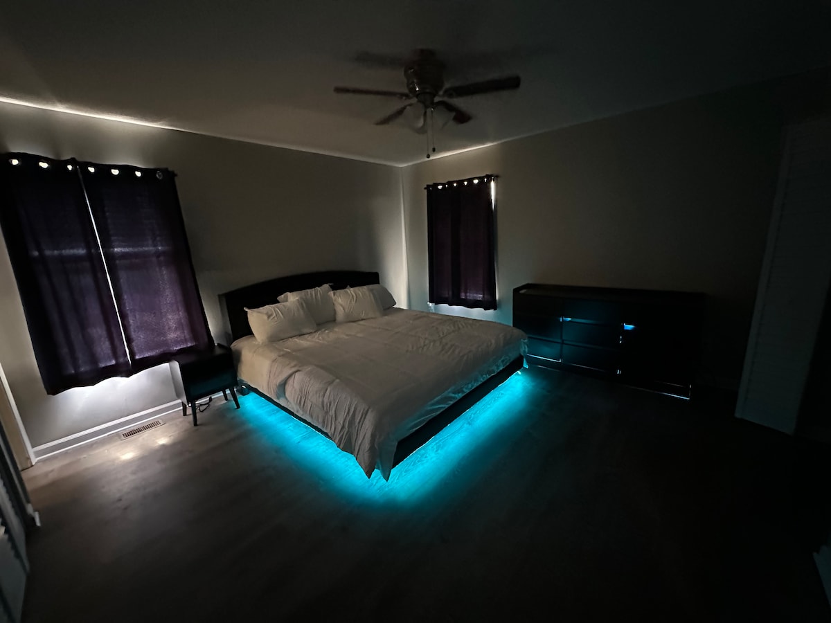 A bedroom is highlighted by a spacious bed outfitted with soft bedding. Ambient lighting around the bed creates a gentle glow. Two dark curtains frame the windows, and a sleek dresser is positioned against one wall, contributing to a contemporary feel.