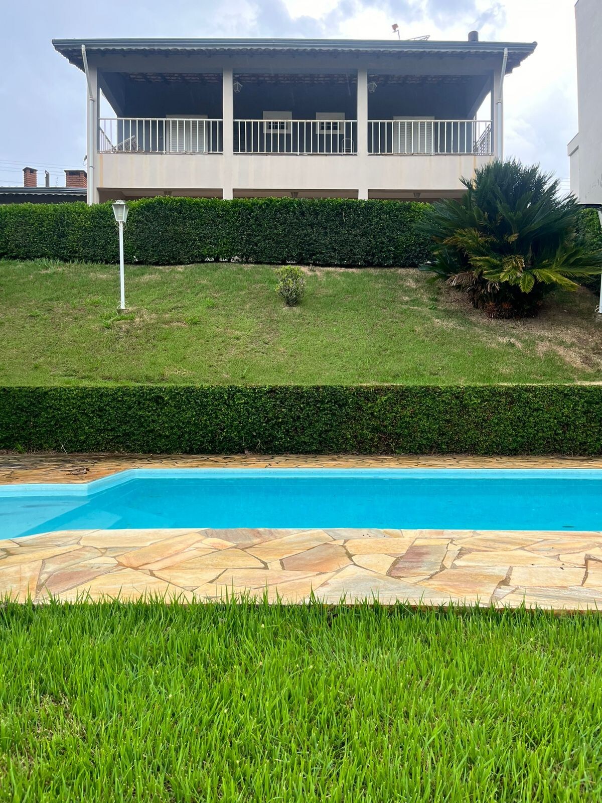 A tranquil outdoor area showcases a clear blue pool, bordered by natural stone. Lush green grass and neatly trimmed hedges frame the pool. A two-story house with a spacious balcony overlooks the scene, offering a serene view of the surrounding landscape.