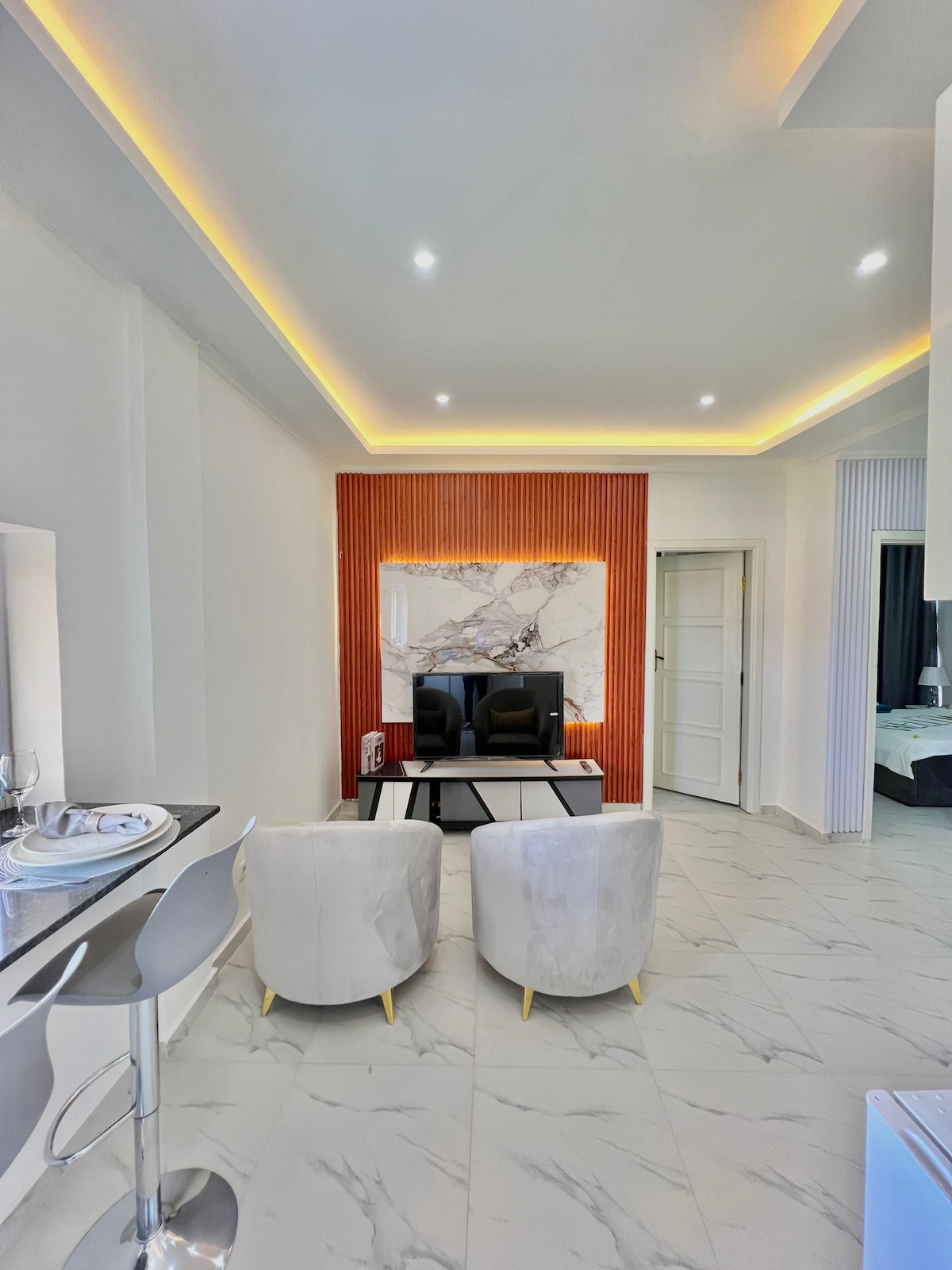A modern living area is highlighted by a marble accent wall and warm lighting. Two plush chairs in light fabric are positioned across from a sleek coffee table, creating a cozy seating arrangement. A glimpse of the bedroom can be seen through an open door.