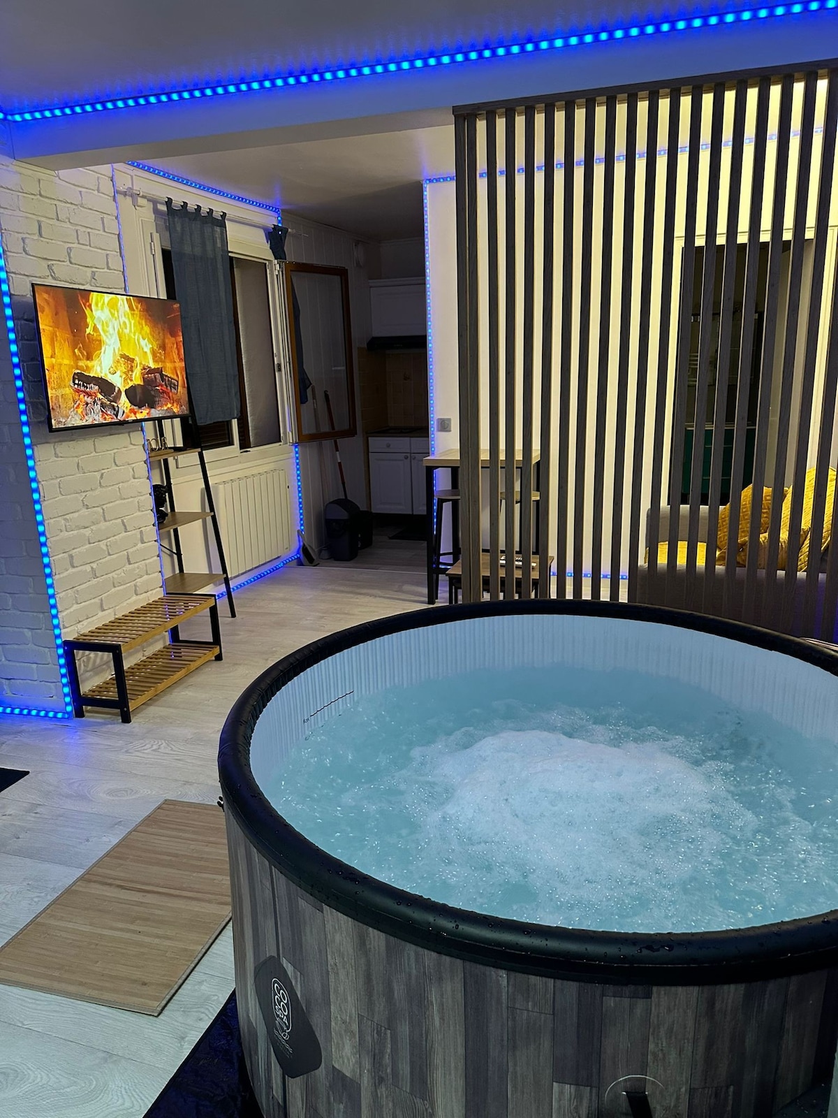A wooden bubble spa is centrally positioned, featuring bubbling water. A television mounted on an adjacent wall displays a warm fire scene. LED lighting creates a soothing ambiance along the walls, enhancing the space, while a cozy yellow sofa is visible in the background.