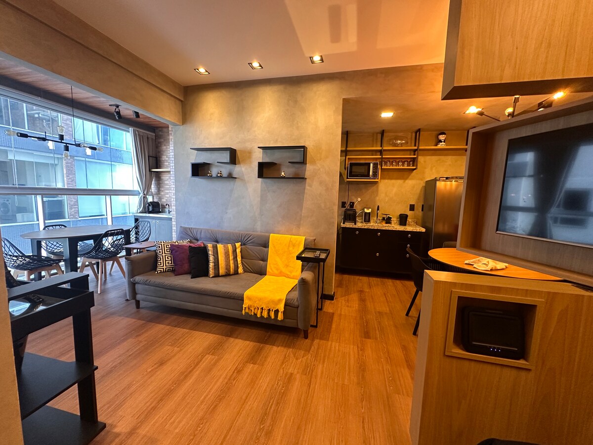 An open-concept living area features a modern sofa adorned with a yellow throw blanket. Dark wooden cabinets and countertops complement the kitchen area, which includes a microwave and an integrated refrigerator. A dining table with six chairs is positioned near large windows, allowing natural light to fill the space.