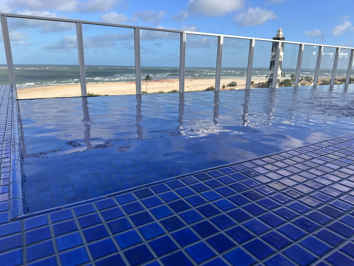 An infinity pool with vibrant blue tiles extends towards the horizon, offering views of the beach and ocean. A lighthouse can be seen in the distance, framed by a clear sky featuring scattered clouds.