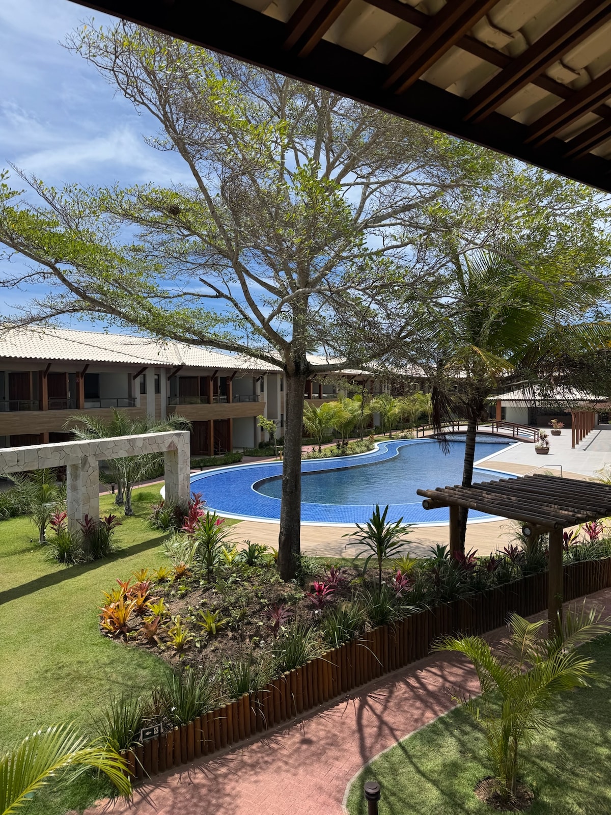 A view of the outdoor area shows a large swimming pool surrounded by tropical plants and palm trees. Spacious buildings can be seen in the background, contributing to a serene environment. A pathway lined with greenery leads towards the pool area.