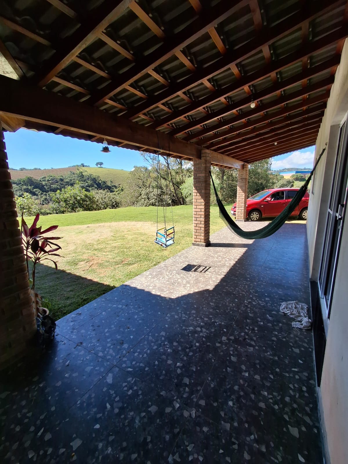 An outdoor porch area features a hammock hanging between two columns and a view of a grassy landscape. A red vehicle is parked nearby. Natural light shines through, casting shadows on the textured floor, while a swing is visible in the distance.
