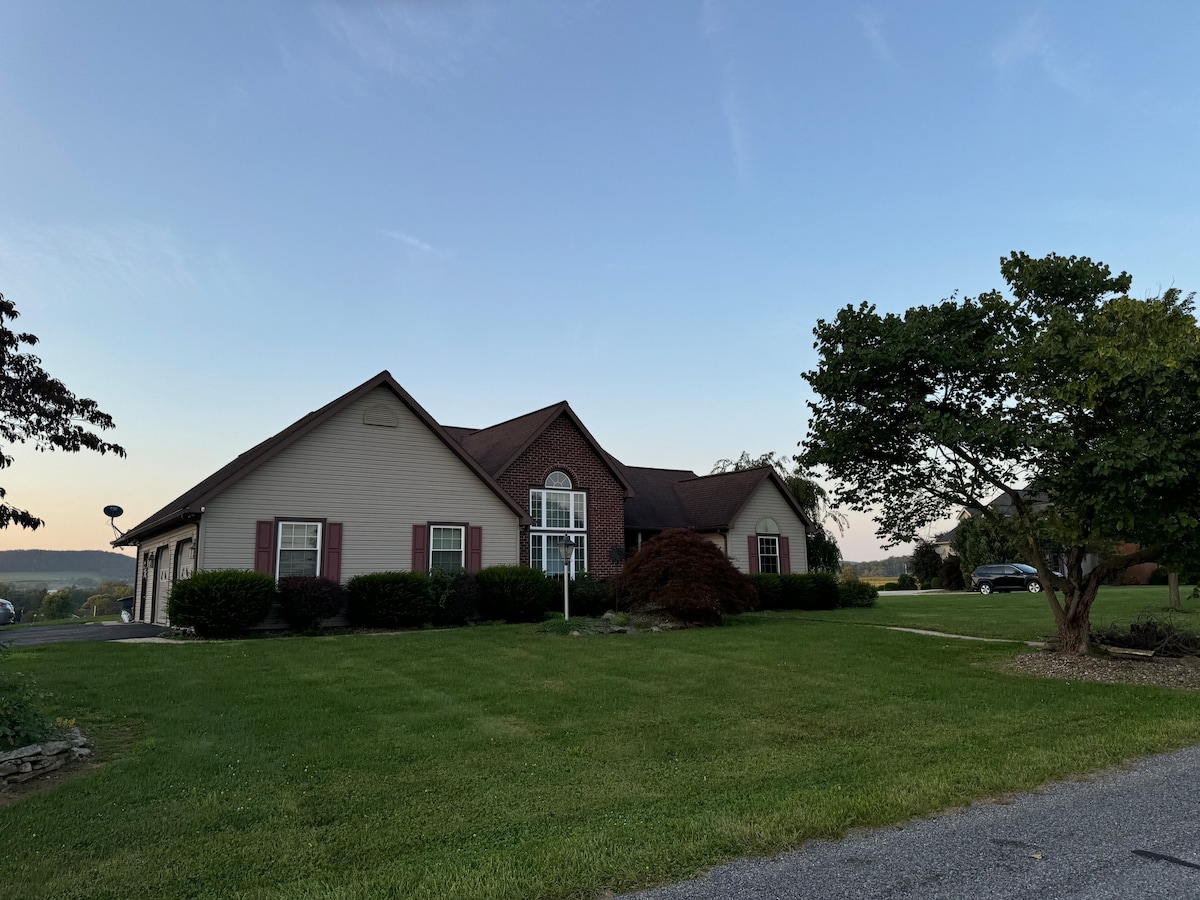 A single-story house is positioned on a grassy hillside, framed by neatly trimmed shrubs. Its inviting facade features light-colored siding with dark roof shingles and windows, complementing the serene suburban landscape. Parking is available nearby, with a clear view of the distant hills.