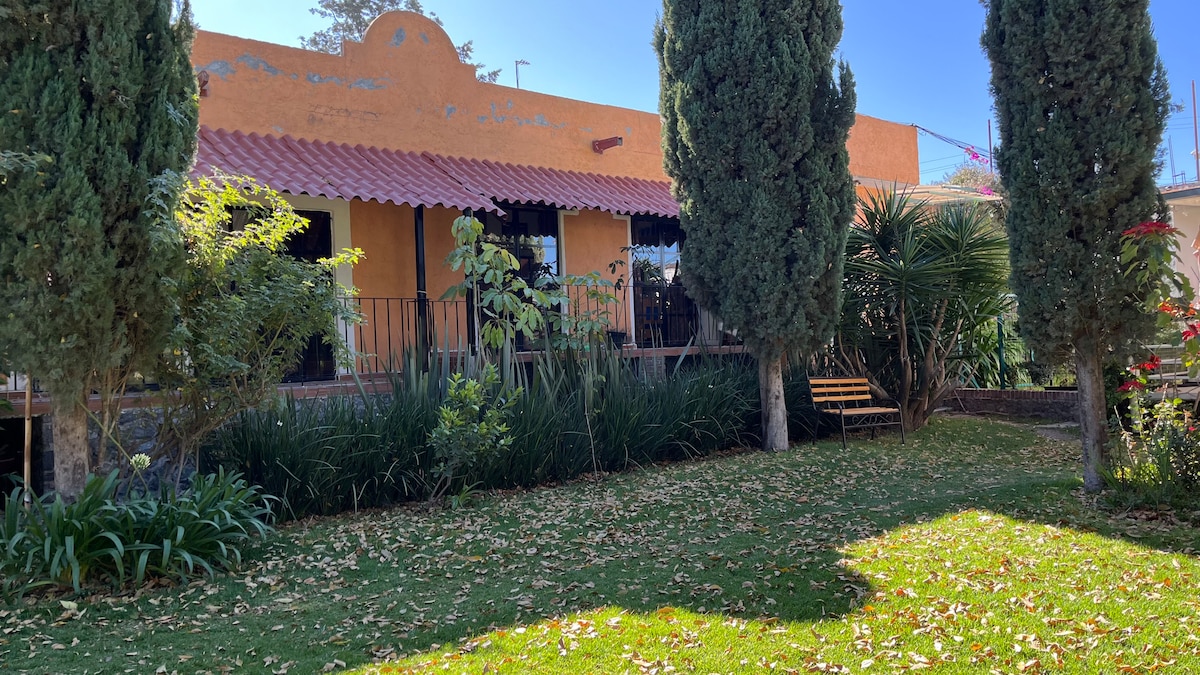 A colonial-style building is framed by neatly maintained greenery and shrubs. A variety of tall trees provide shade over the garden, which features a patch of grass scattered with fallen leaves. The inviting patio area is visible, with seating arranged for guest relaxation.