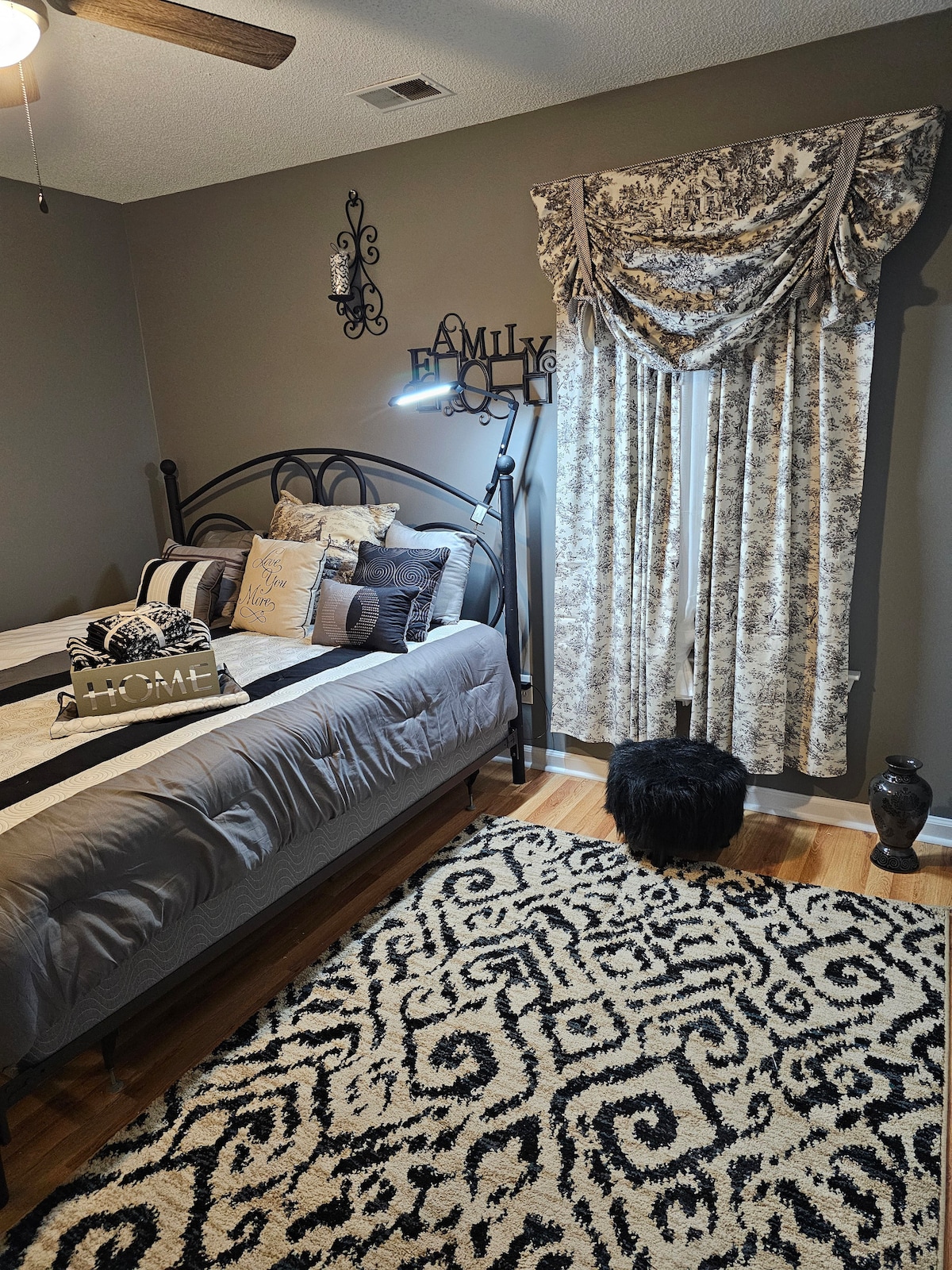 A cozy sleeping area features a decorative bed with an assortment of pillows, accompanied by a stylish lamp. Textured curtains frame the window, while a black and white area rug adds contrast to the hardwood floor. A small decorative vase and an accent piece are situated nearby.