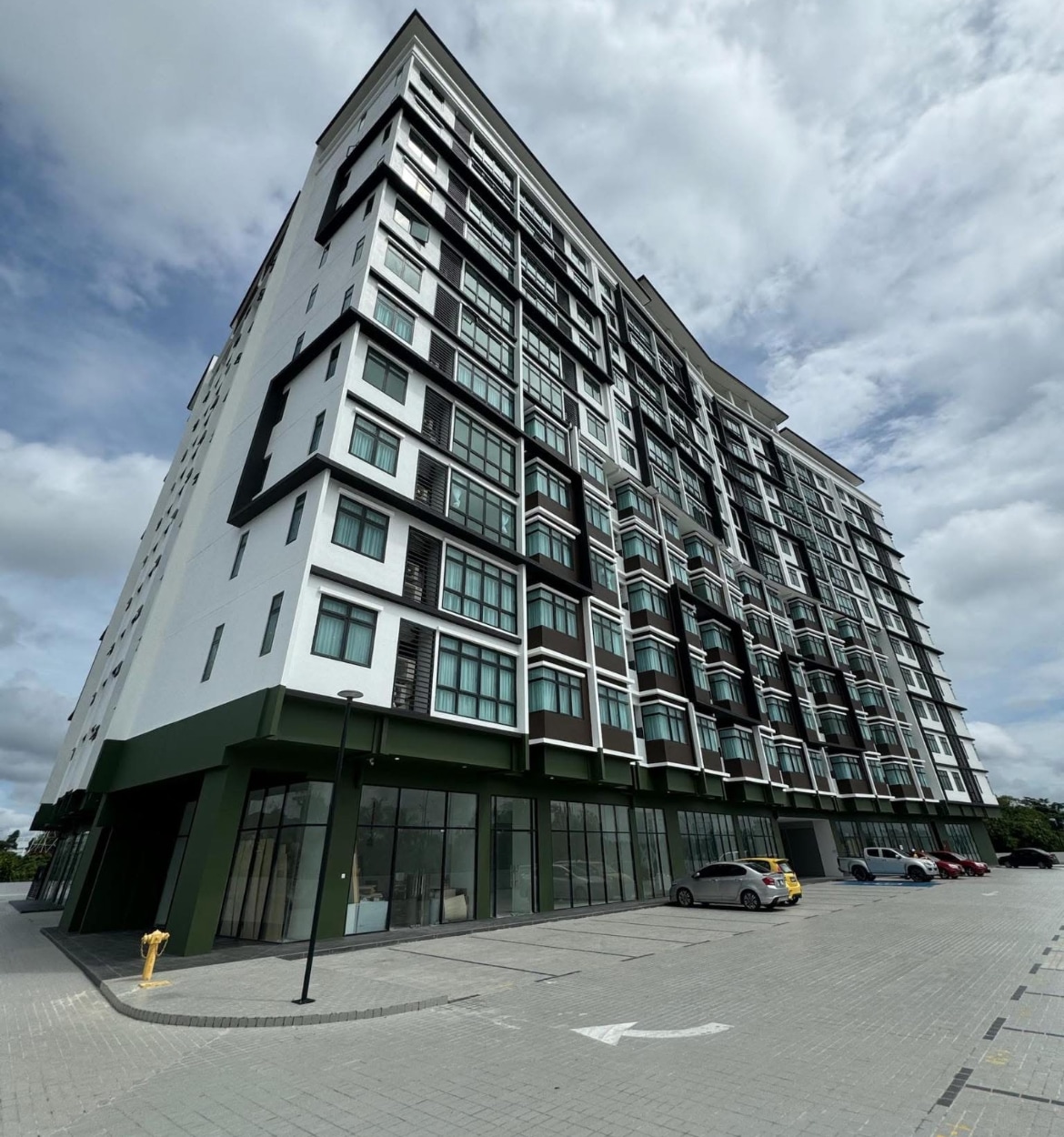 A modern multi-story building is displayed, featuring large windows and a sleek design. The structure is complemented by a spacious parking area with neatly arranged vehicles. A cloudy sky casts a gentle light over the scene, enhancing the building's contemporary appearance.