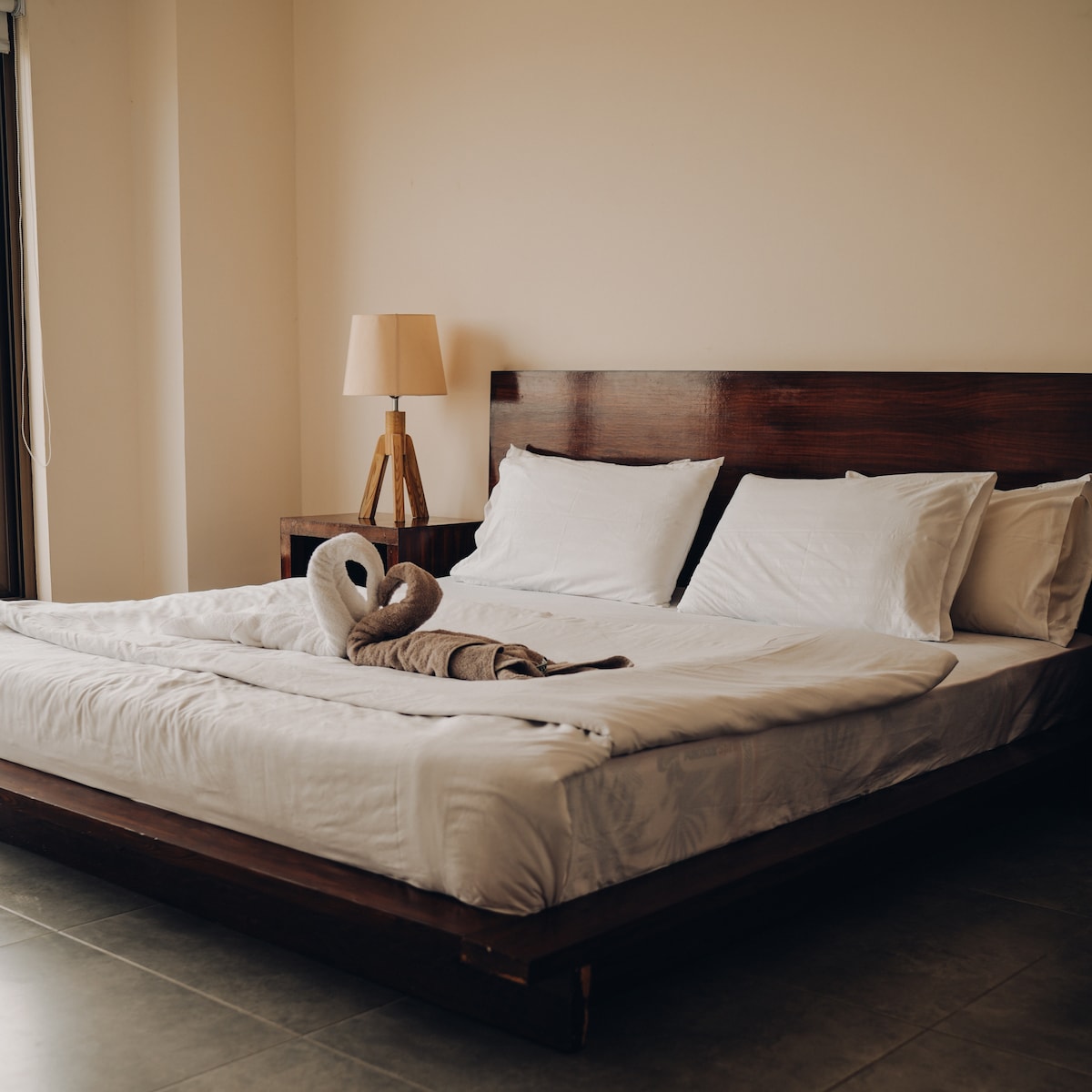 A spacious bedroom features a large wooden bed with crisp white linens and multiple pillows. A wooden nightstand holds a stylish lamp, while natural light filters through a nearby window, enhancing the calm ambiance of the room.