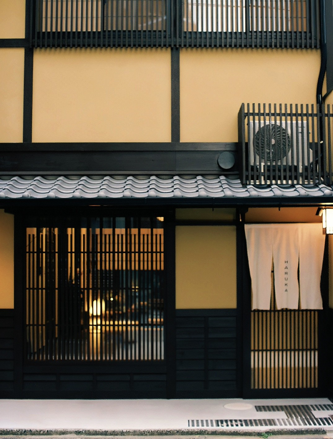 The exterior of a two-story traditional townhome is presented, featuring wooden slats and a tiled roof. The entrance is adorned with a fabric sign and soft lighting from within creates a welcoming ambiance, while an air conditioning unit is discreetly located to the side.
