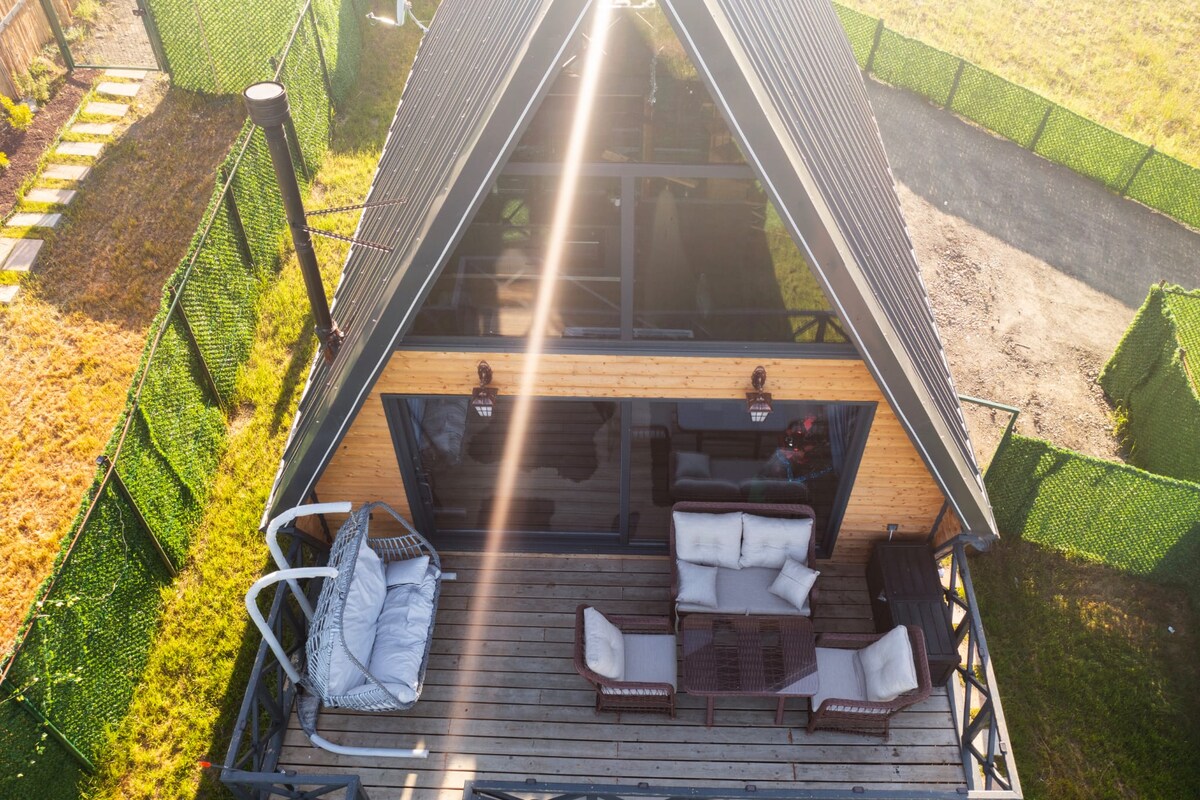 An aerial view showcases a cozy wooden cabin with a sloped roof. A spacious outdoor deck is furnished with comfortable seating, including a swing chair and a seating arrangement, surrounded by a green privacy fence and open natural surroundings.
