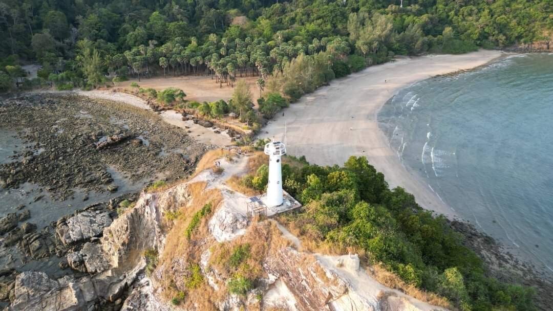 An aerial view captures a solitary lighthouse perched on a rocky outcrop, overlooking a serene beach and clear waters. Lush greenery lines the coastline, contrasting with the sandy shore. The tranquil scene is complemented by gentle waves lapping at the edge of the beach.