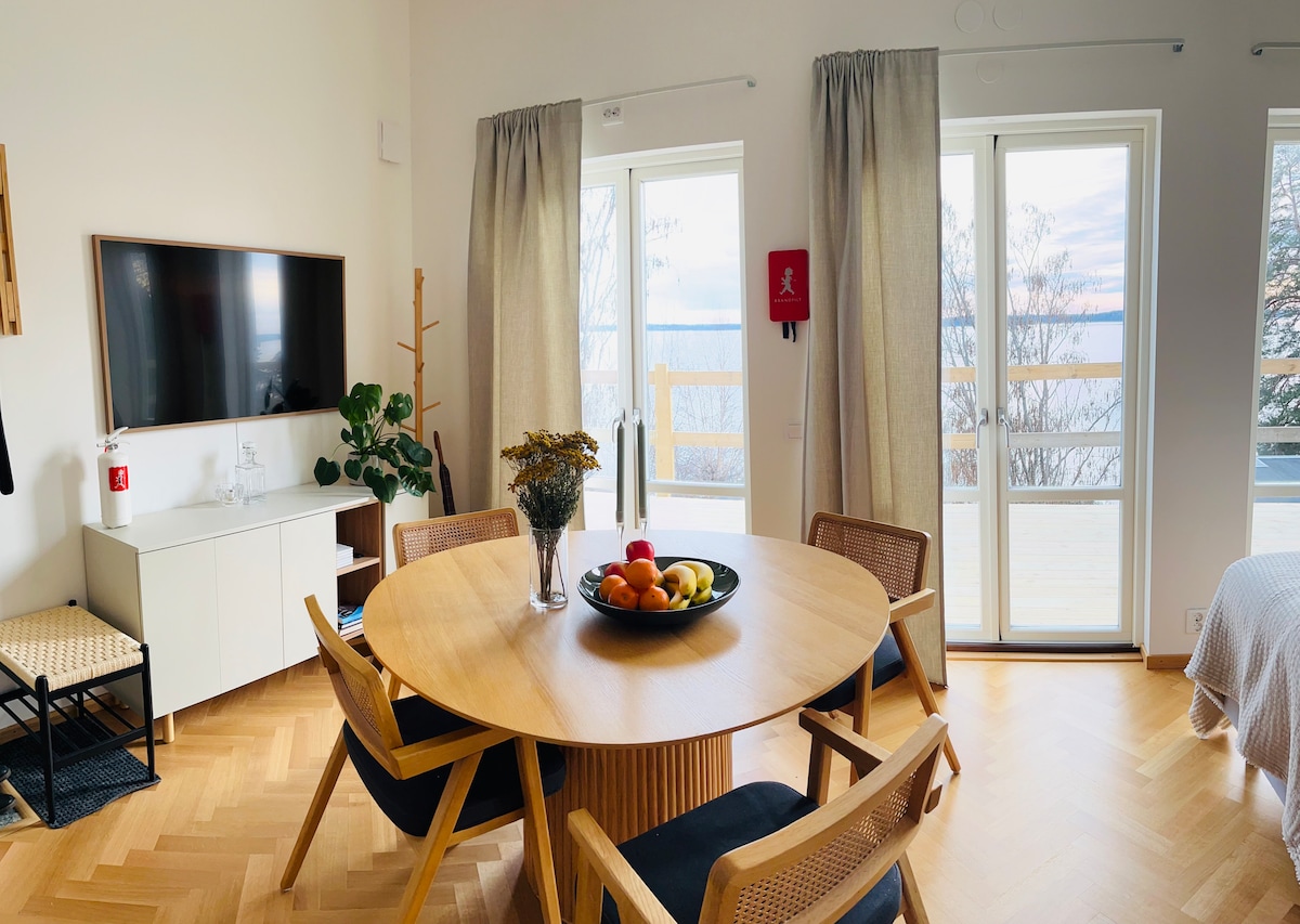 A round wooden dining table is surrounded by four wooden chairs. A bowl of fresh fruit is placed at the center. Large glass doors provide views of the outdoor area, allowing natural light to brighten the open space, which features a flat-screen television mounted on the wall.