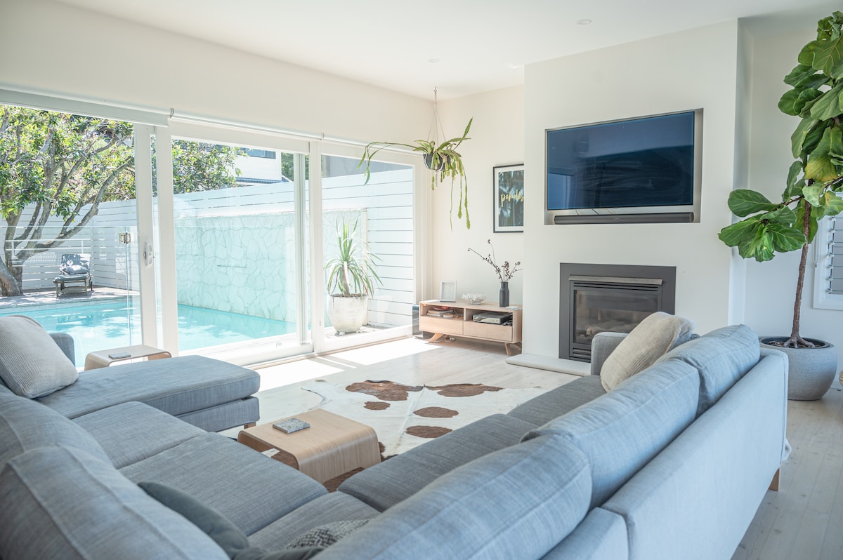 An open-plan living area is showcased with a large sectional couch facing a wall-mounted television. Natural light floods the space through expansive glass doors that offer a view of the private magnesium pool and surrounding greenery.