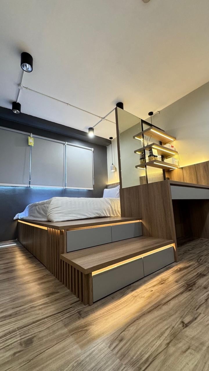 A modern bedroom features a raised sleeping area accessible by steps, illuminated with soft lighting. A workstation is integrated into the design, with shelves mounted against the wall. Large windows allow natural light to fill the space, complemented by neutral-toned walls.