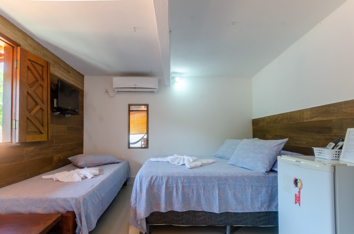 A tranquil sleeping area is displayed with two spacious beds adorned with light blue bedding. A small television is mounted on the wall, and a white refrigerator sits in the corner. Natural light streams through a wooden window with a decorative shutter.