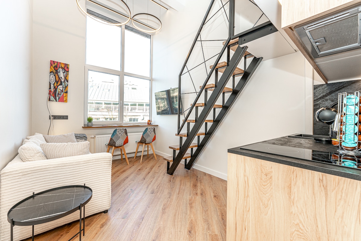 A modern living space features a comfortable sofa and a sleek glass coffee table. Large windows allow natural light to fill the room, while a staircase with wooden steps leads to an upper level. A flat-screen TV is mounted on the wall, complemented by stylish seating.