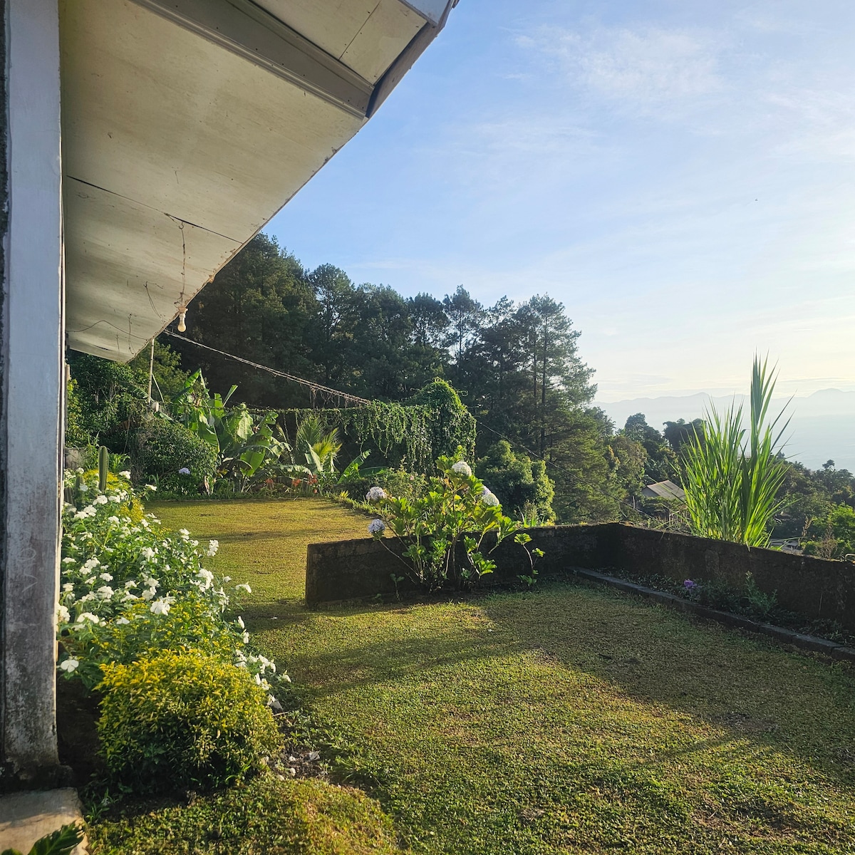 A well-maintained garden area features lush greenery and a variety of flowers, bordered by stone walls. A gentle slope leads to distant mountains, with clear skies illuminating the landscape. Natural light enhances the vibrancy of the outdoor space, ideal for relaxation.