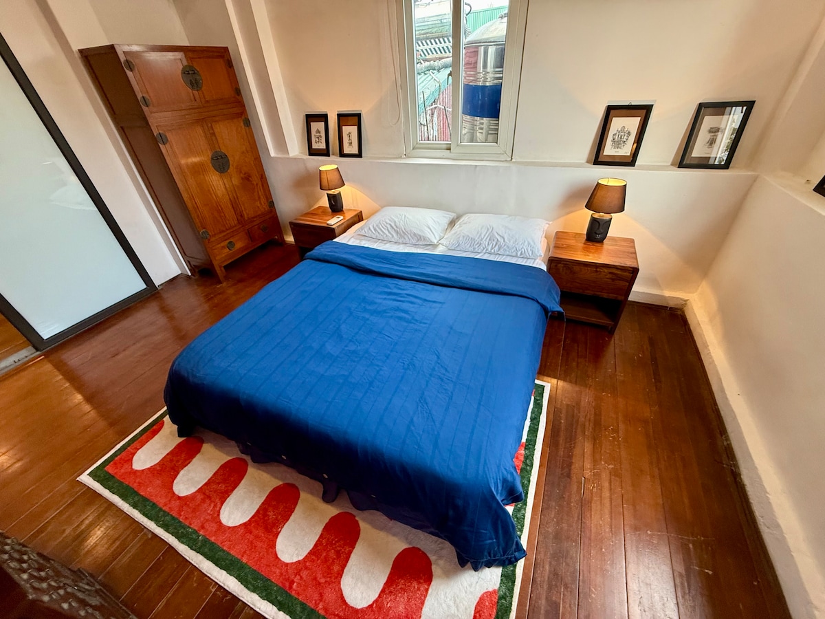 A cozy sleeping area features a spacious bed covered with a blue blanket, flanked by two wooden nightstands with lamps. The room has wooden flooring and is accented by a patterned rug. Framed artwork is displayed on the walls beside the bed.