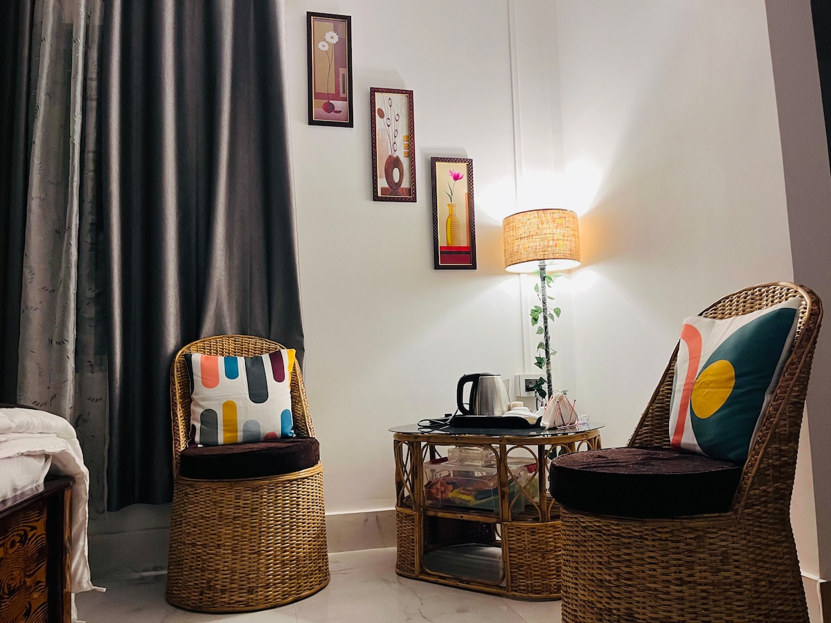 A cozy seating area is presented with two woven chairs, each adorned with decorative cushions. A glass table sits in the center, accompanied by a kettle and various items. The corner lamp provides soft illumination, while framed artworks adorn the wall.