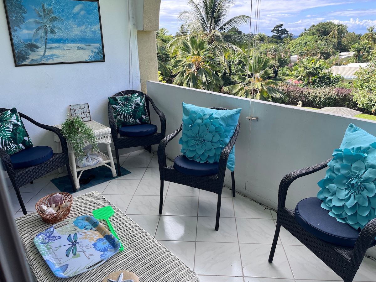 A private balcony is furnished with four wicker chairs, each adorned with bright blue and green cushions. A small table with decorative items sits in the center. Lush tropical foliage and a clear sky create a serene backdrop, providing a calming environment for relaxation.