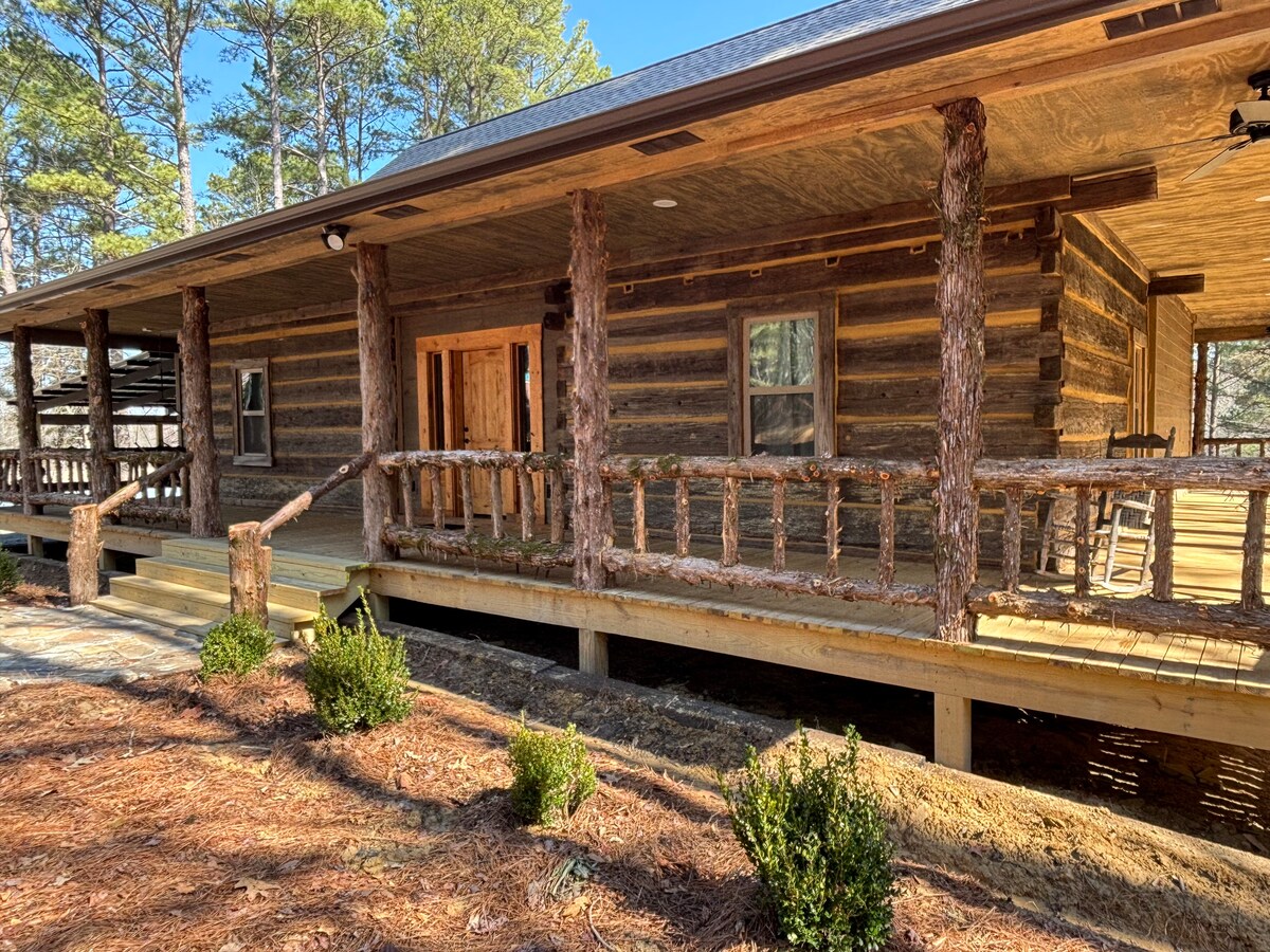 The log house features a spacious porch with wooden railings, providing a natural and rustic aesthetic. The exterior showcases traditional log construction, framed by surrounding trees and landscaping. The entrance is accessible via a gradual ramp leading up to the porch.