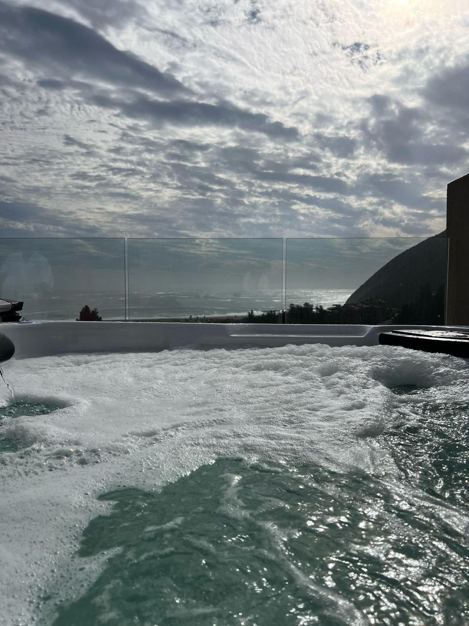A jacuzzi is featured on a terrace, with foam and bubbles visible on the surface of the water. A panoramic view of the sea and clouds stretches in the background, framed by a clear glass railing that ensures unobstructed sightlines.