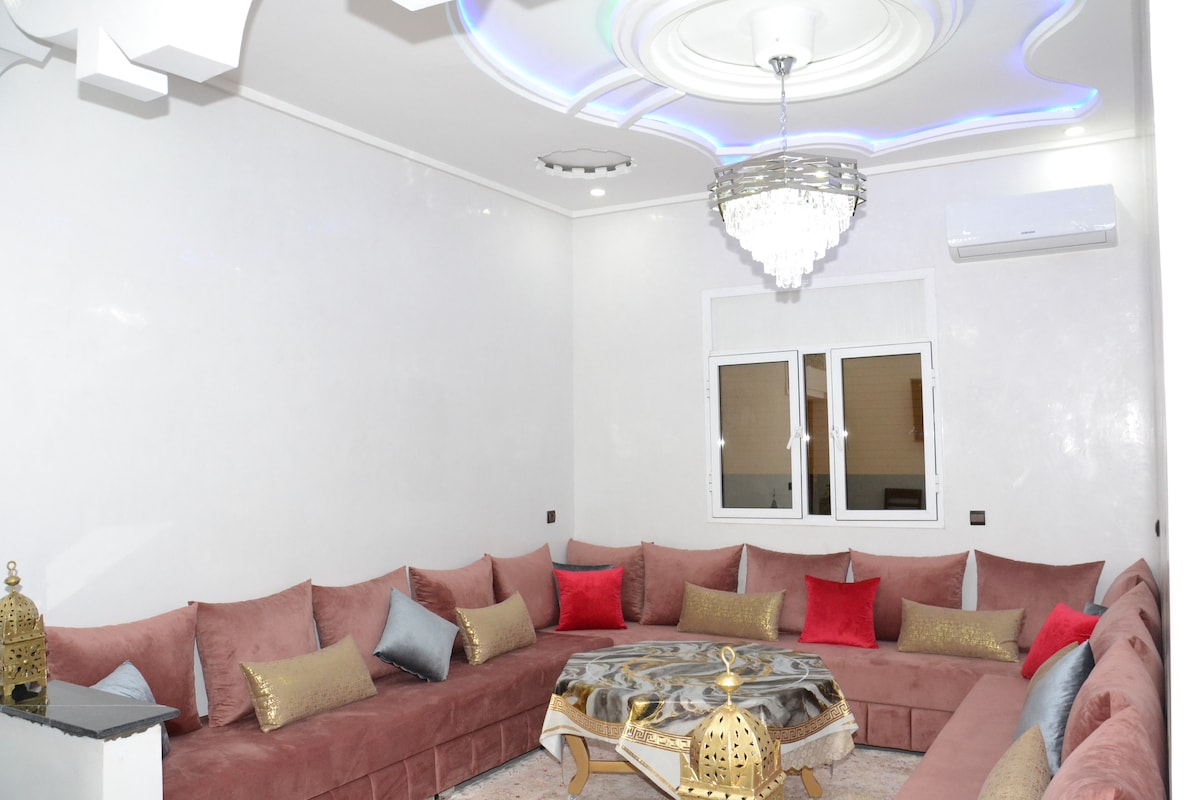 A spacious seating area features a large U-shaped sofa adorned with colorful cushions, complemented by a decorative coffee table at the center. Elegant chandelier lighting enhances the soft ambiance, while white walls and a modern ceiling design contribute to the overall aesthetic.
