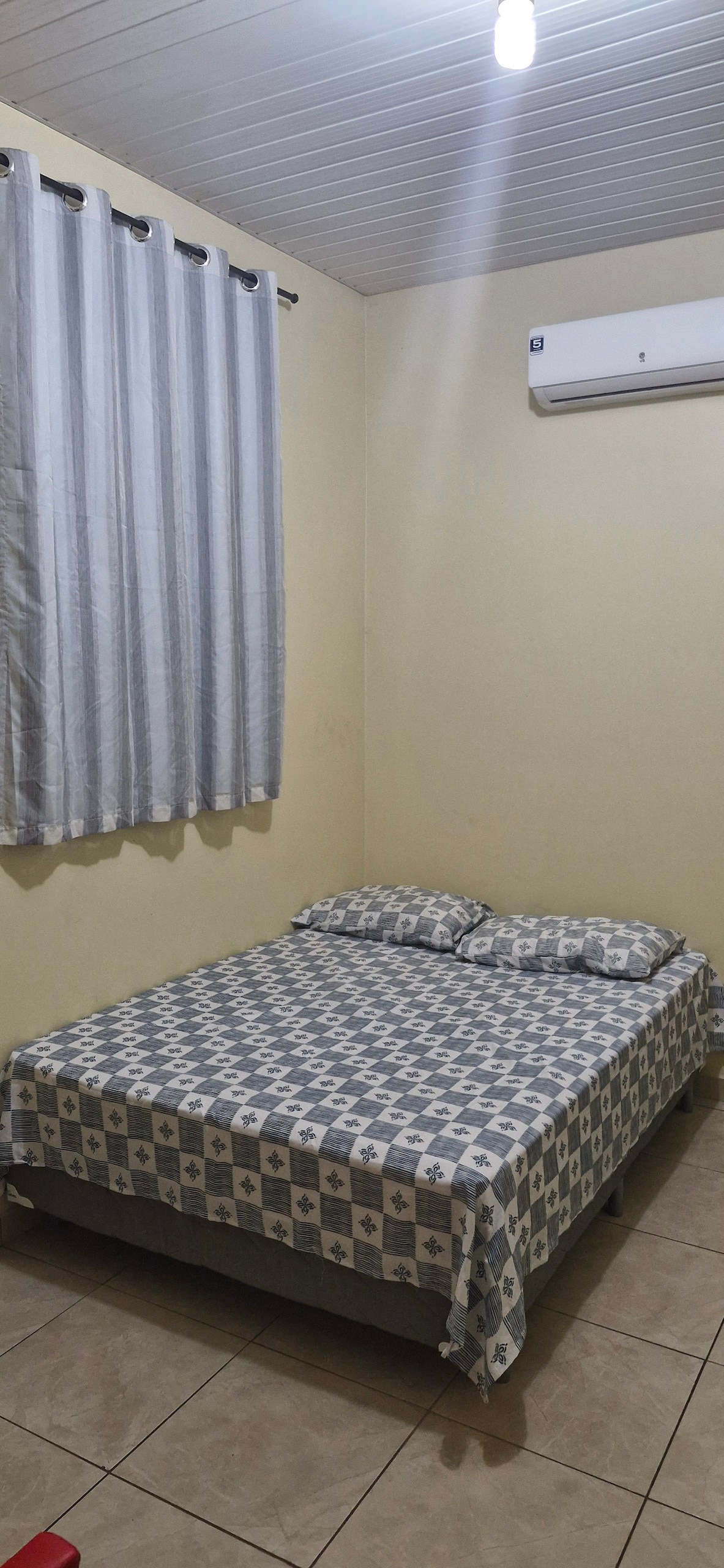 A bedroom is presented with a spacious bed covered in a patterned blanket and positioned against a light-colored wall. Neutral tiles line the floor, while a window dressed with sheer curtains allows natural light to illuminate the space. An air conditioning unit is mounted above.