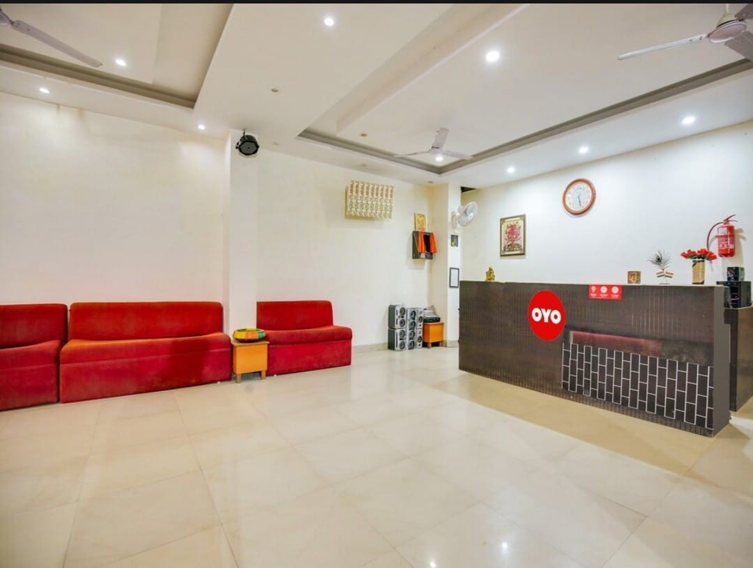 A welcoming lobby area features a reception desk with modern detailing. Two red sofas provide seating options, complemented by light-colored walls and a neutral tiled floor. A wall clock and framed artwork add subtle decor, while ceiling fans enhance comfort in the open space.