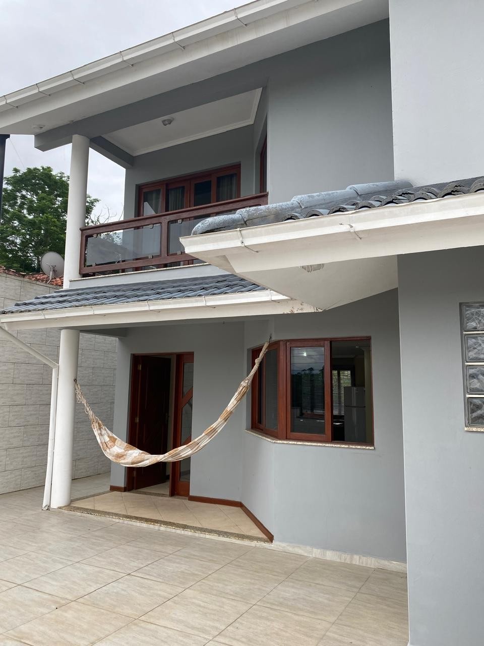 A two-story house displays a light gray exterior with balconies above the entrance. A hammock is hung in a spacious patio, which is paved with large tiles. Windows framed in dark wood offer access to natural light, enhancing the welcoming atmosphere.
