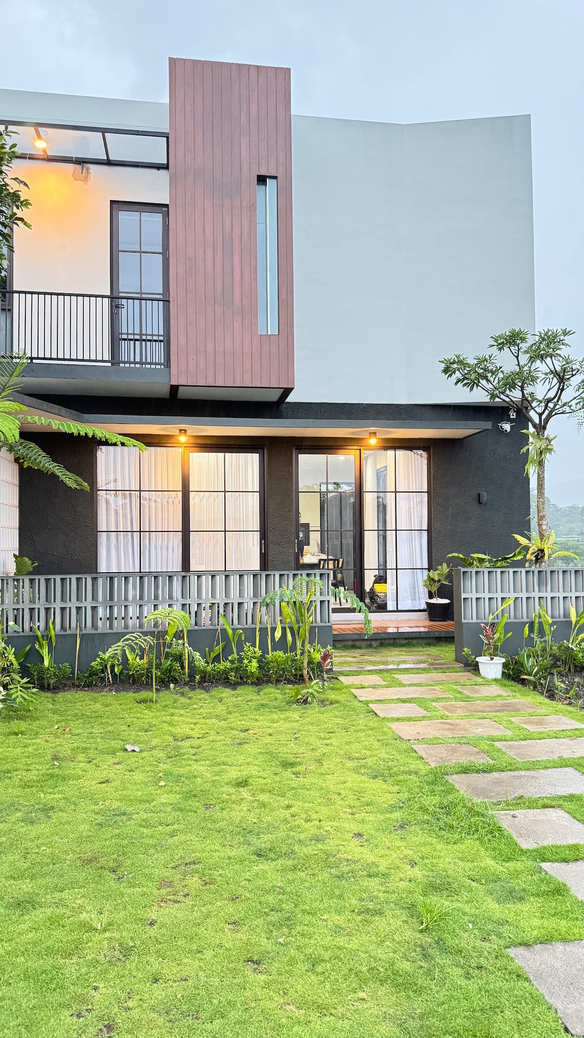 The villa exterior is surrounded by a lush garden, featuring a neatly manicured lawn and a stone pathway. Large windows and a porch area are visible, allowing for ample natural light. Soft lighting highlights the modern architectural elements of the building.