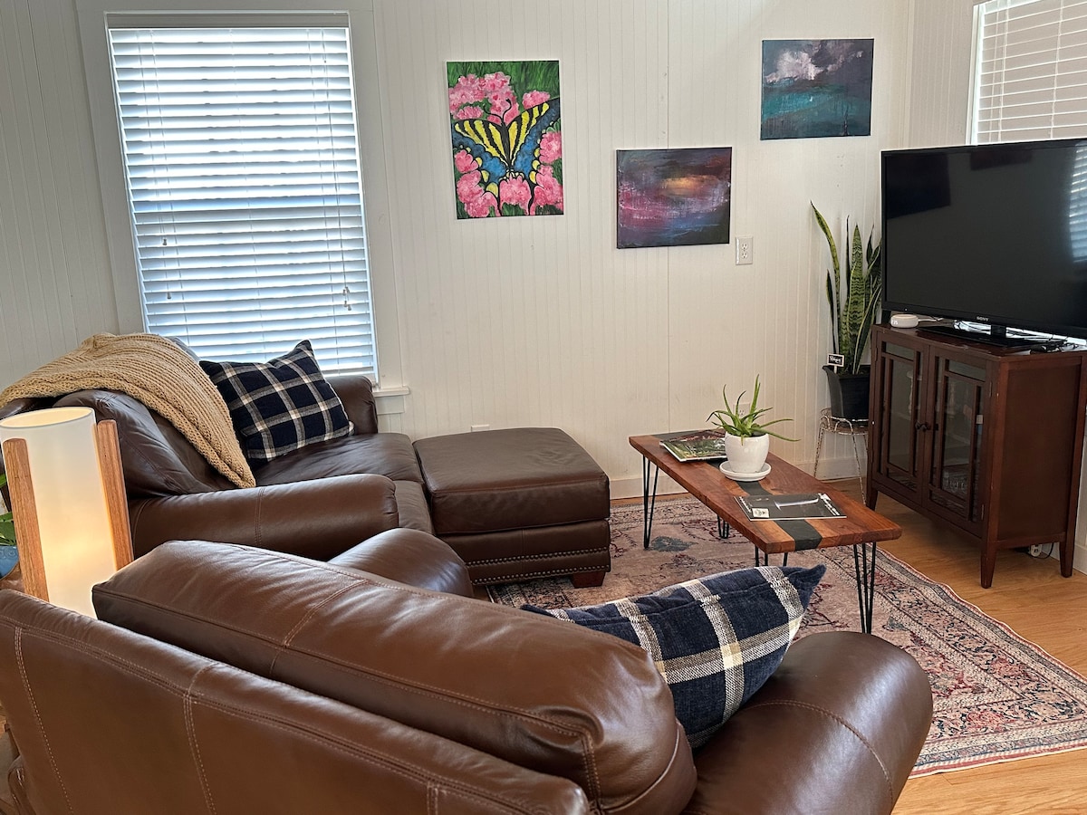 A living room is presented with a brown leather sectional sofa and a matching armchair. Colorful artwork adorns the walls, and a television is positioned opposite a wooden coffee table. A plant is placed on the table, contributing to the cozy ambiance.