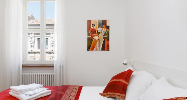 Ljust och modernt sovrum, med vita väggar, dubbelsäng med röda textilier, öppna garderober och lådor. Stort fönster, luftkonditionering och dekorativa målningar kompletterar miljön.