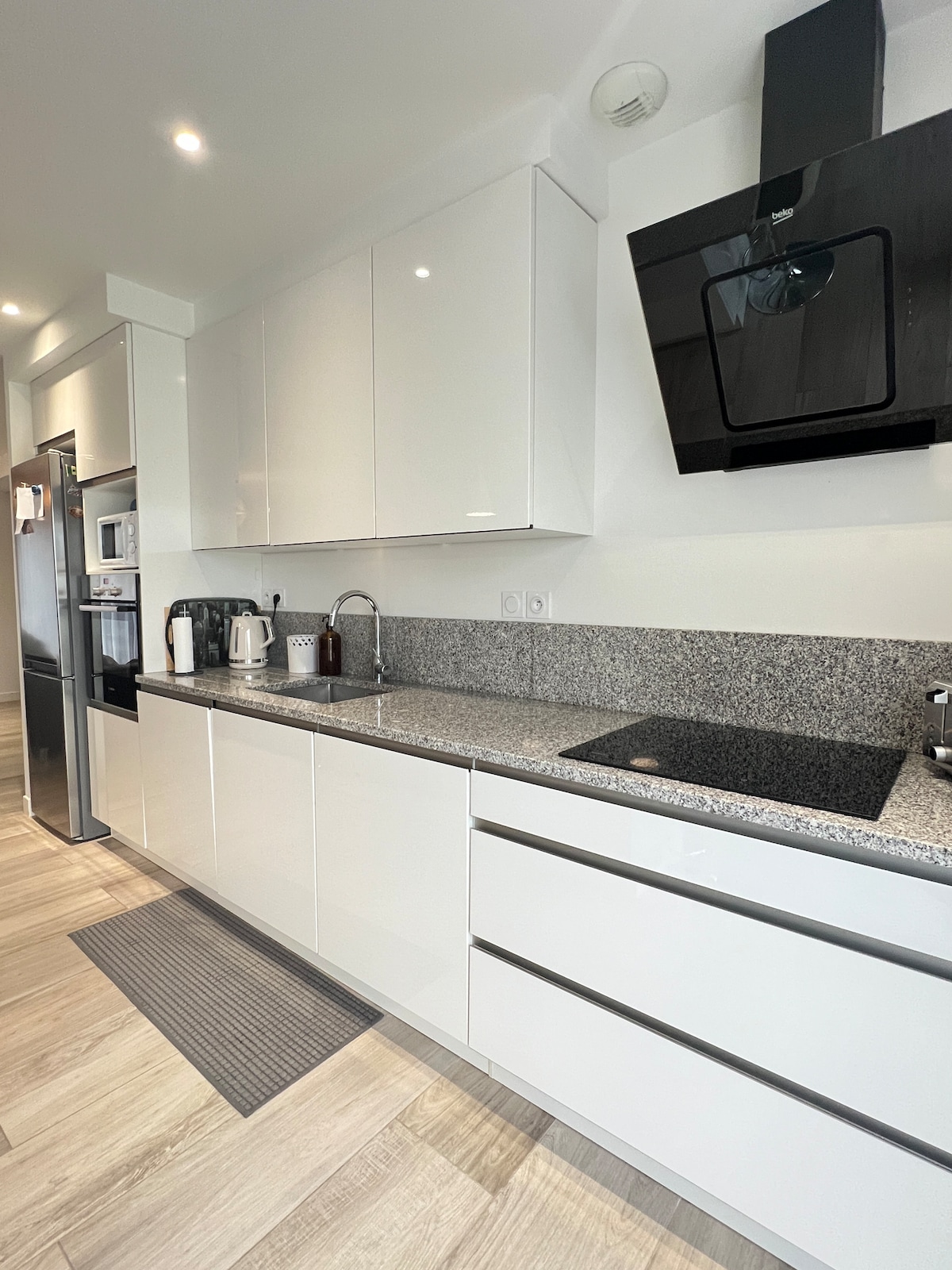 A modern kitchen features sleek white cabinetry and a spacious granite countertop. Essential appliances, including a kettle and coffee maker, are visible. A cooktop is integrated into the design, contributing to the overall contemporary feel of the kitchen space.