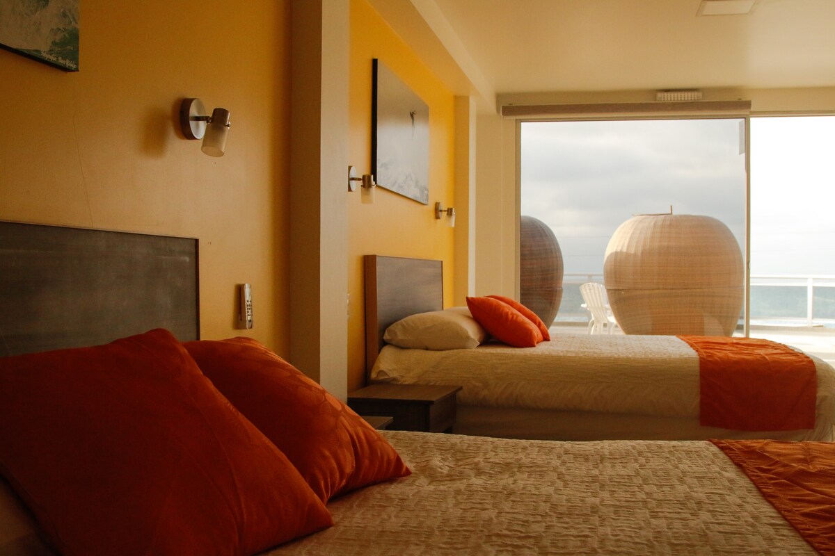 The bright room features two spacious beds dressed in neutral bedding with orange accent pillows. A large glass door invites natural light and showcases a view of the ocean, while comfortable seating is visible on the balcony beyond.
